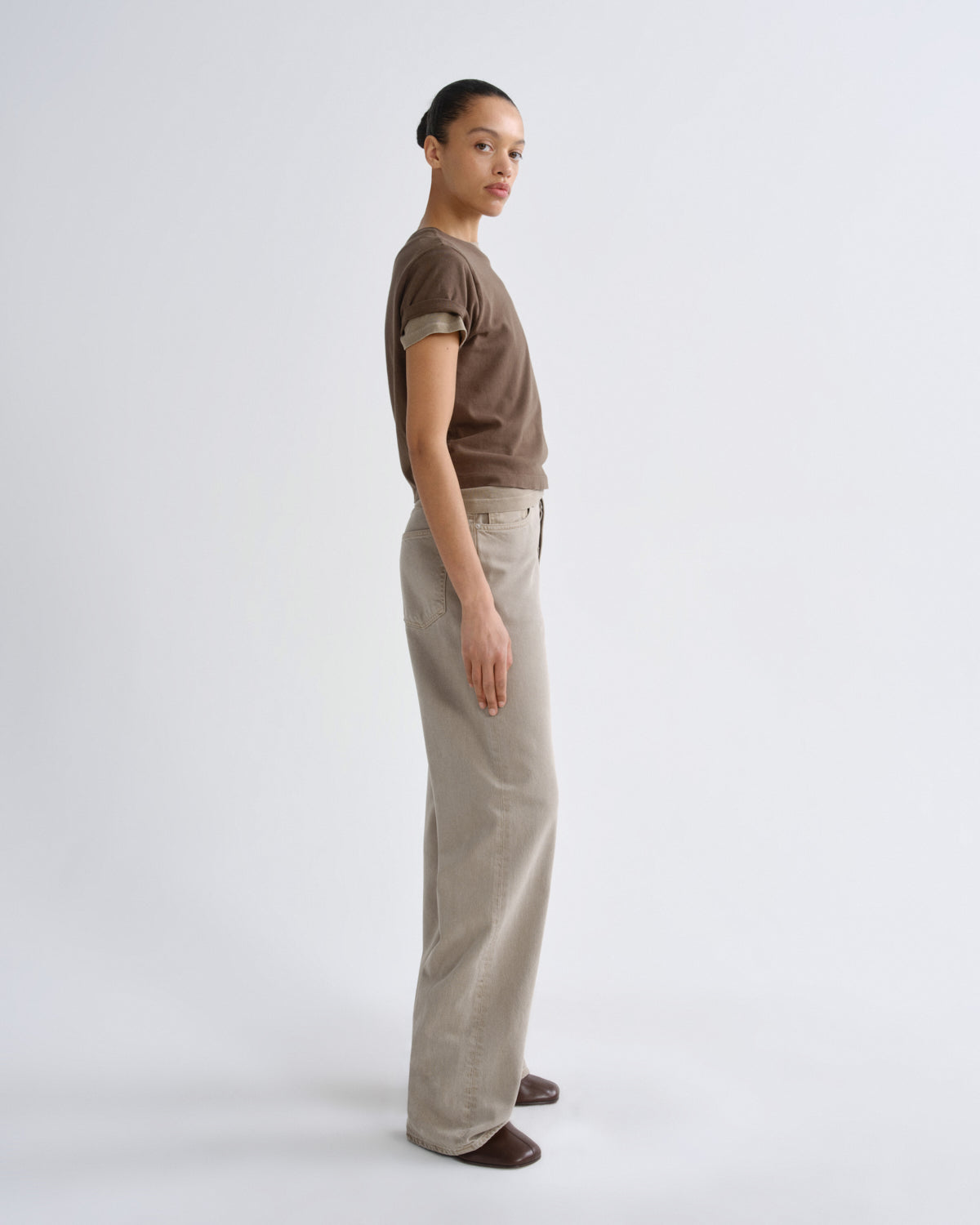 A model stands in profile wearing a brown short-sleeve t-shirt and light beige wide-leg trousers against a plain light gray studio background.