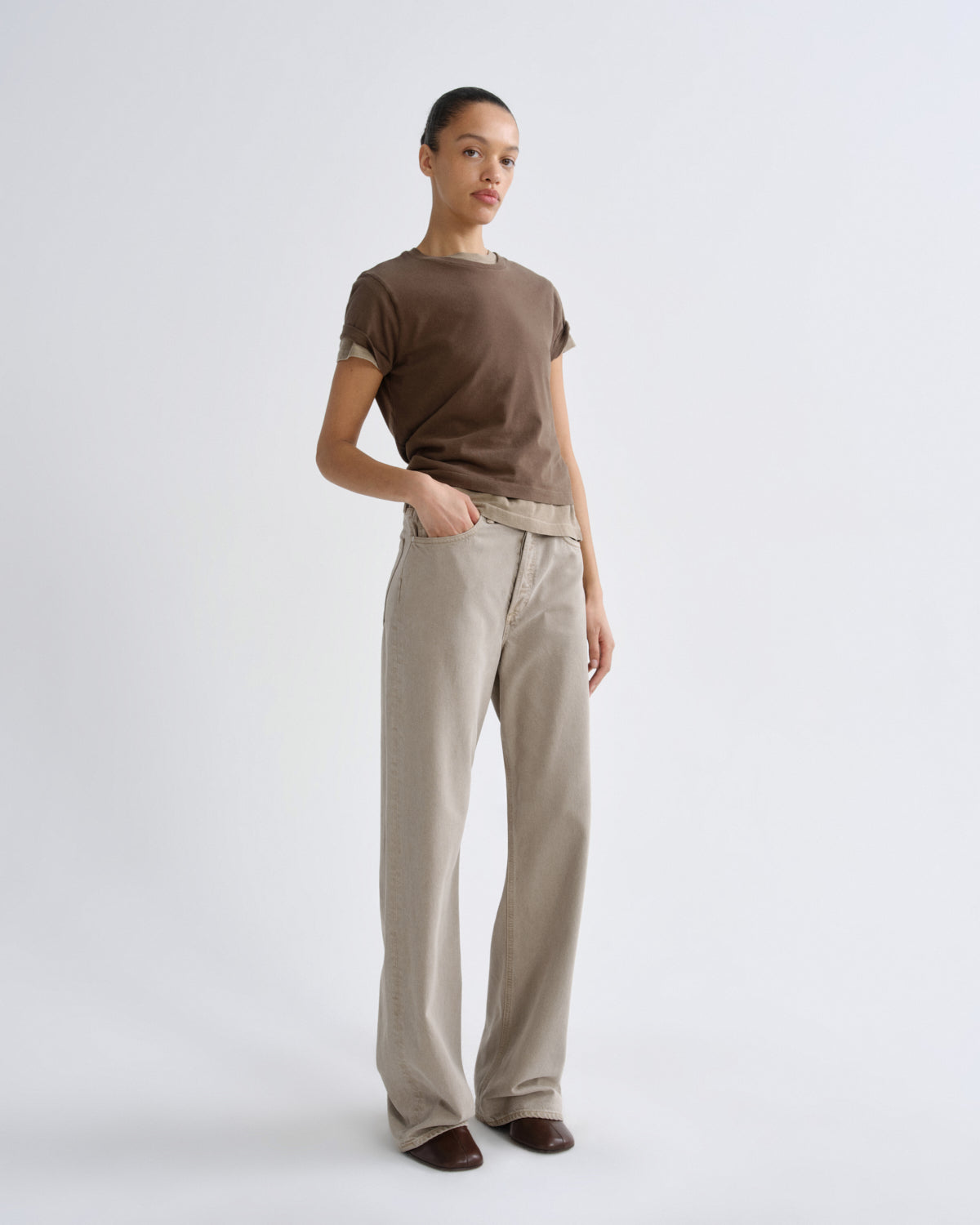 A model stands against a light gray studio background wearing a brown short-sleeve t-shirt and beige wide-leg trousers.