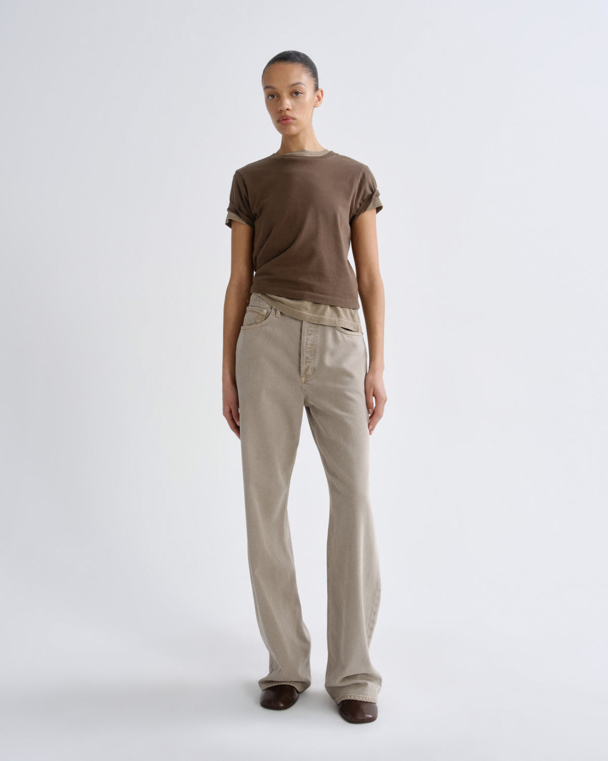 A model stands against a light gray studio background wearing a brown short-sleeve crewneck t-shirt and light beige straight-leg jeans.