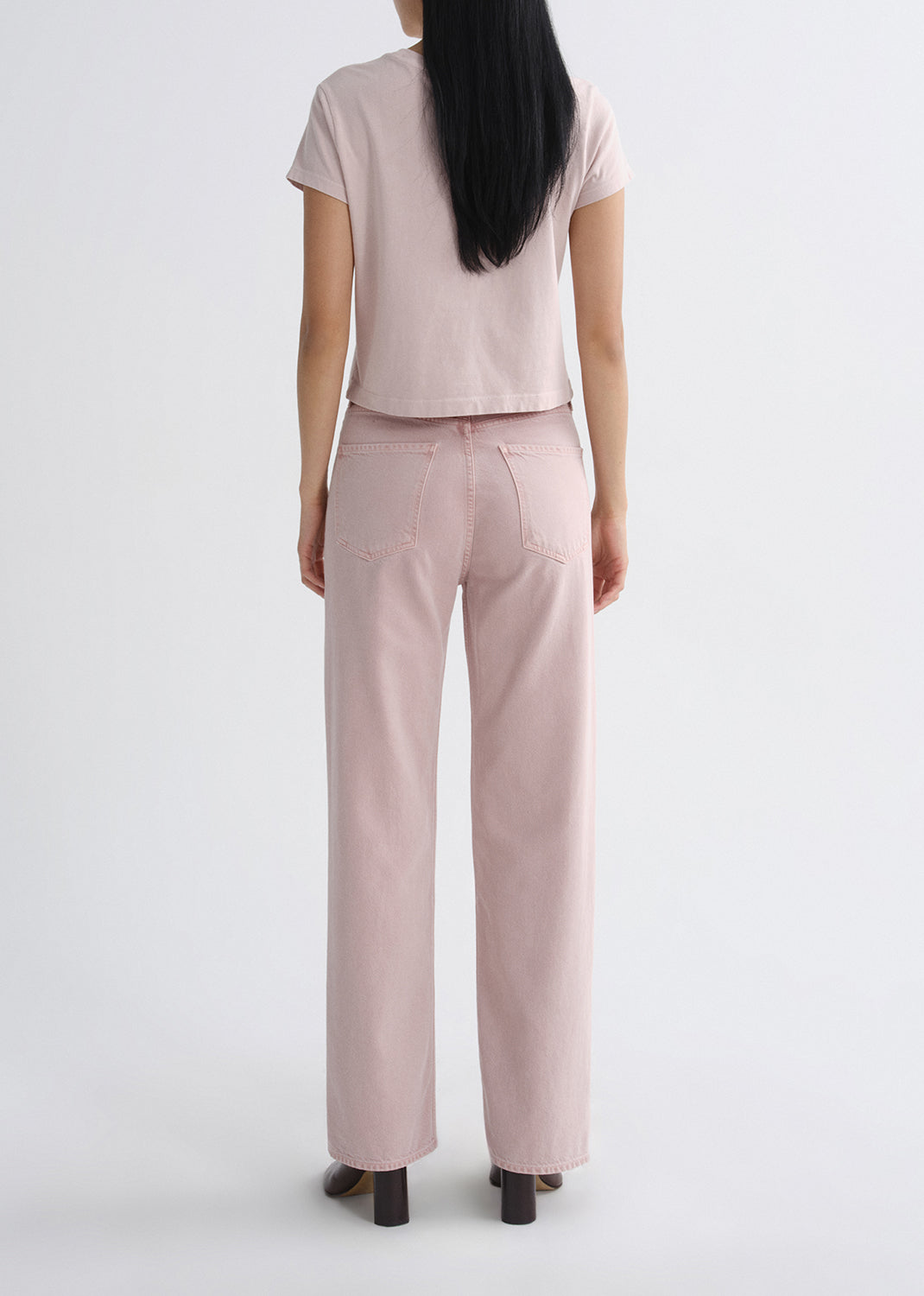 Person wearing a light pink short sleeve crewneck tee with light pink straight leg jeans viewed from the back on a white background