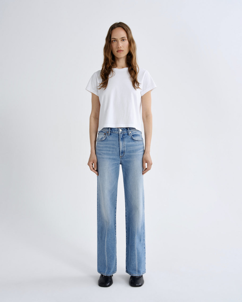 A woman stands against a light gray studio background wearing a white short-sleeve cropped T-shirt, light-wash blue wide-leg jeans, and black shoes.