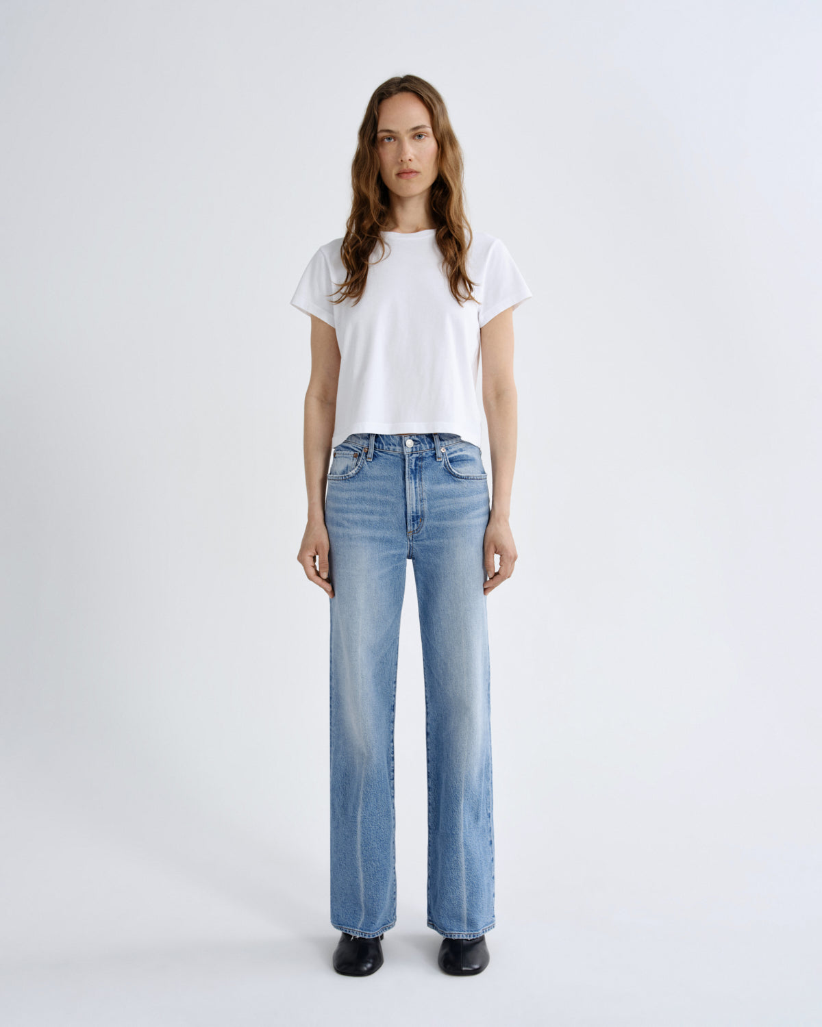 A woman stands against a light gray studio background wearing a white short-sleeve cropped T-shirt, light-wash blue wide-leg jeans, and black shoes.