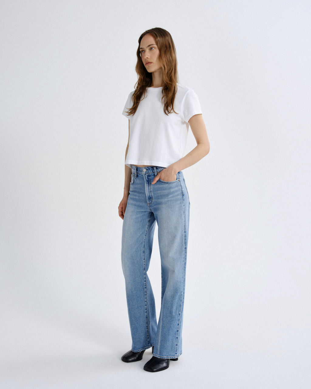 A woman modeling a white short-sleeve crop top and light-wash blue straight-leg jeans against a plain light gray studio background.