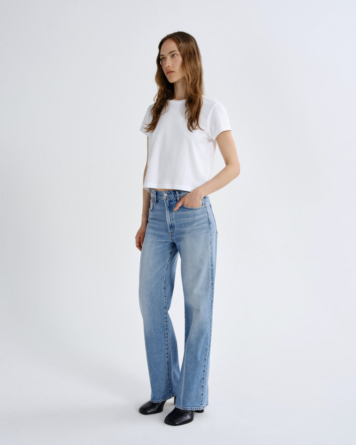 A woman modeling a white short-sleeve crop top and light-wash blue straight-leg jeans against a plain light gray studio background.