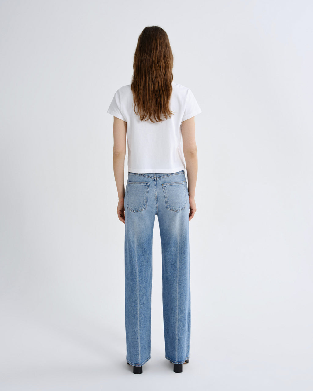 A woman viewed from behind wearing a white short-sleeve t-shirt and light-wash wide-leg blue jeans, standing against a plain light gray studio background.