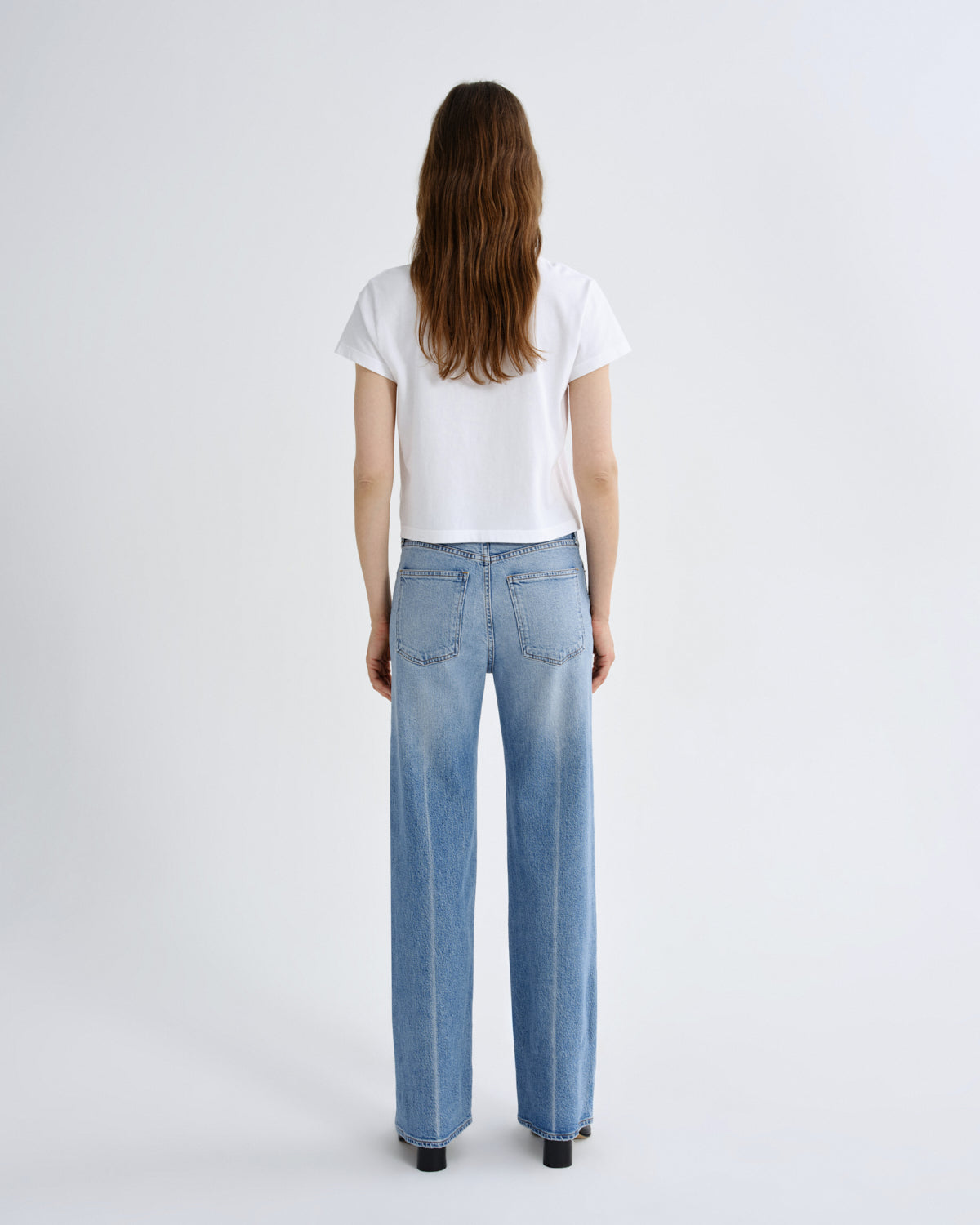 A woman viewed from behind wearing a white short-sleeve t-shirt and light-wash wide-leg blue jeans, standing against a plain light gray studio background.