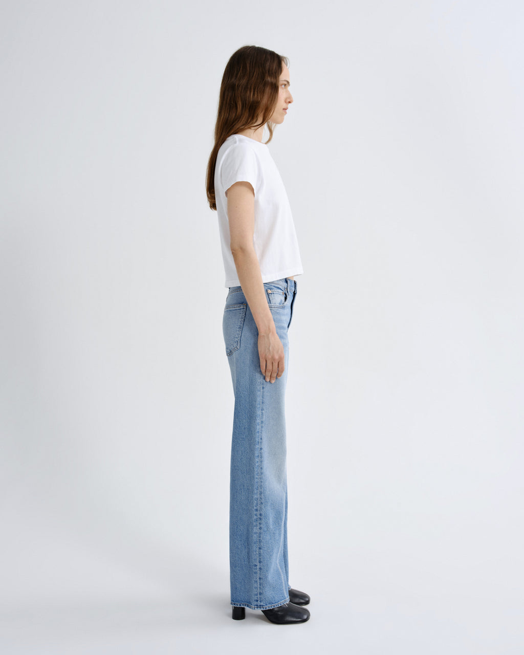 A woman in a white short-sleeve t-shirt and light-wash wide-leg blue jeans stands in profile against a clean, light gray studio background.