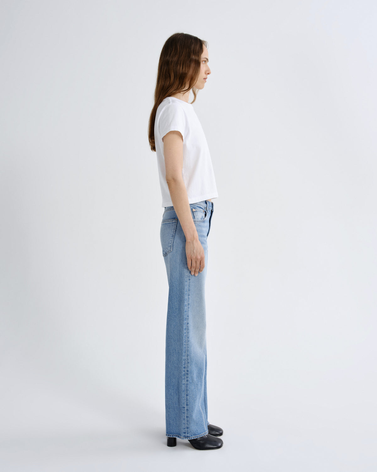 A woman in a white short-sleeve t-shirt and light-wash wide-leg blue jeans stands in profile against a clean, light gray studio background.