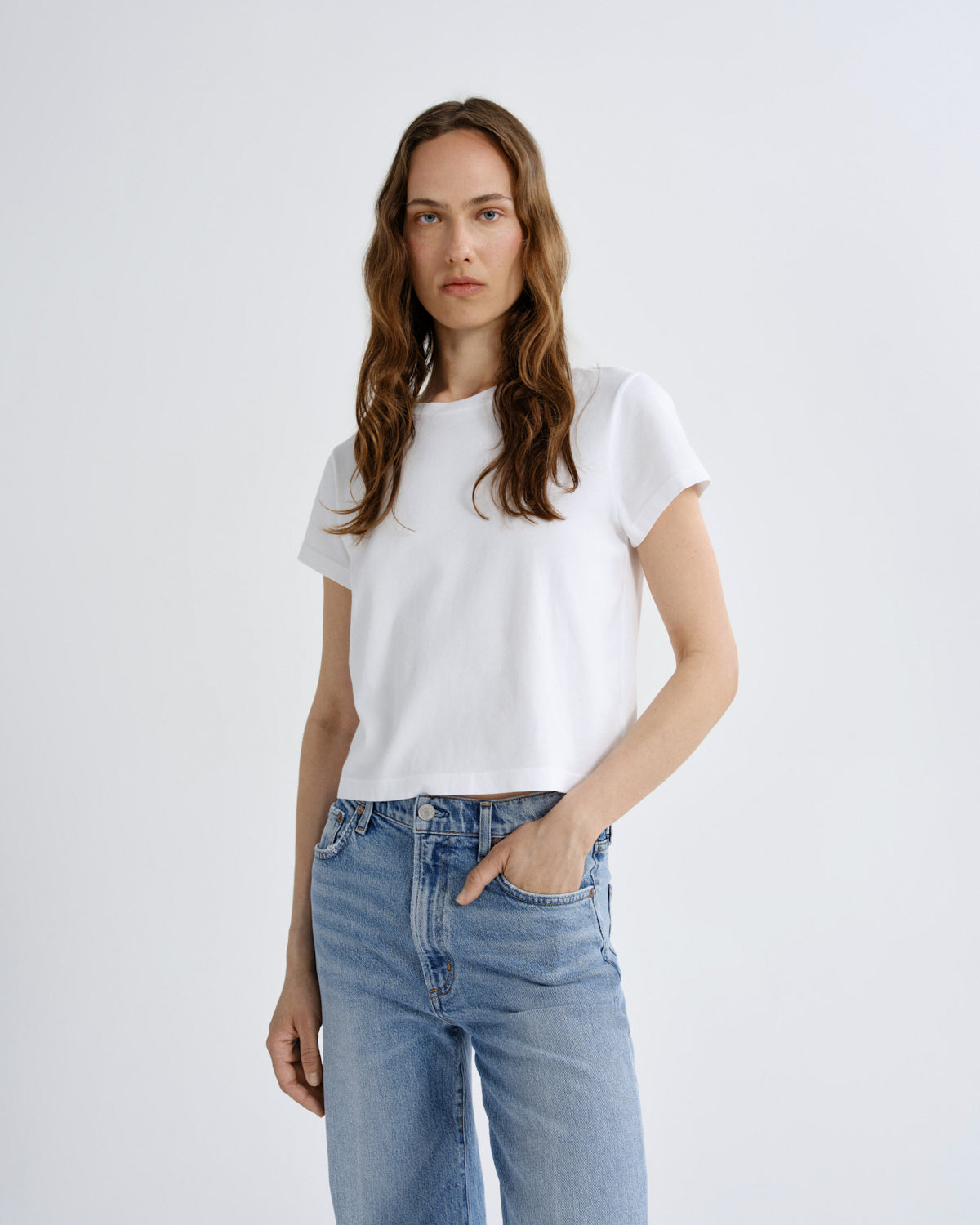 A woman modeling a white short-sleeve crewneck t-shirt paired with light-wash blue jeans, standing against a plain light gray studio background.