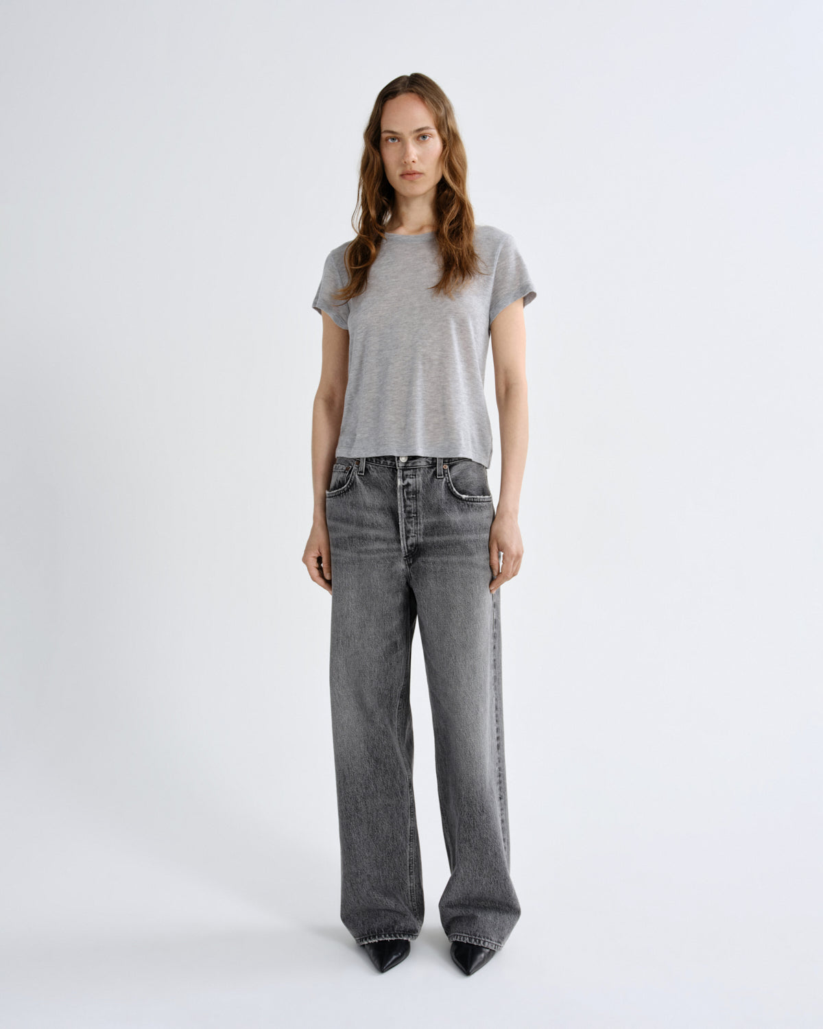 A woman stands against a light gray studio background wearing a light gray short-sleeve t-shirt, wide-leg faded gray jeans, and black pointed shoes.
