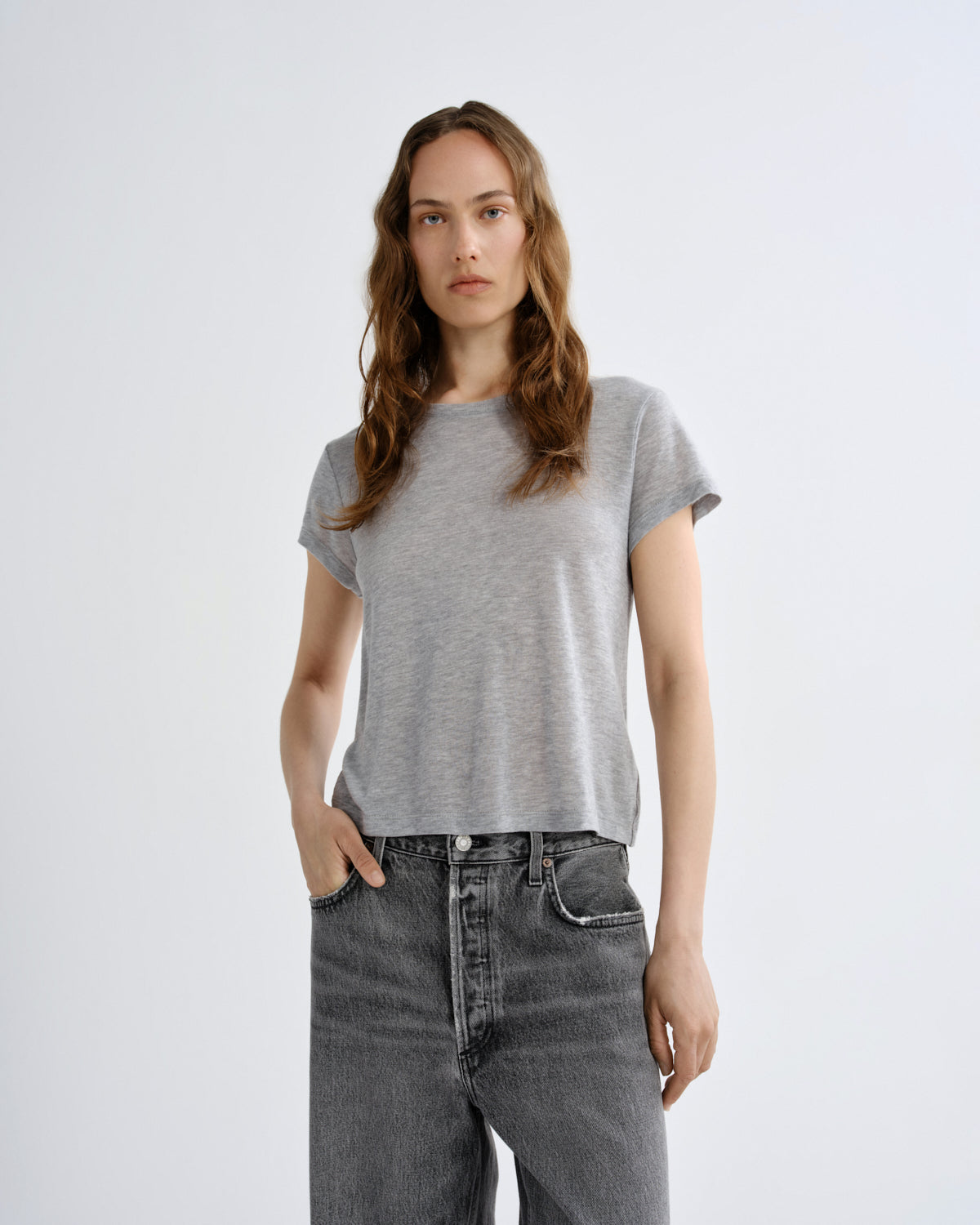A woman modeling a light gray short-sleeve crewneck t-shirt paired with dark gray jeans, standing against a plain light gray studio background.