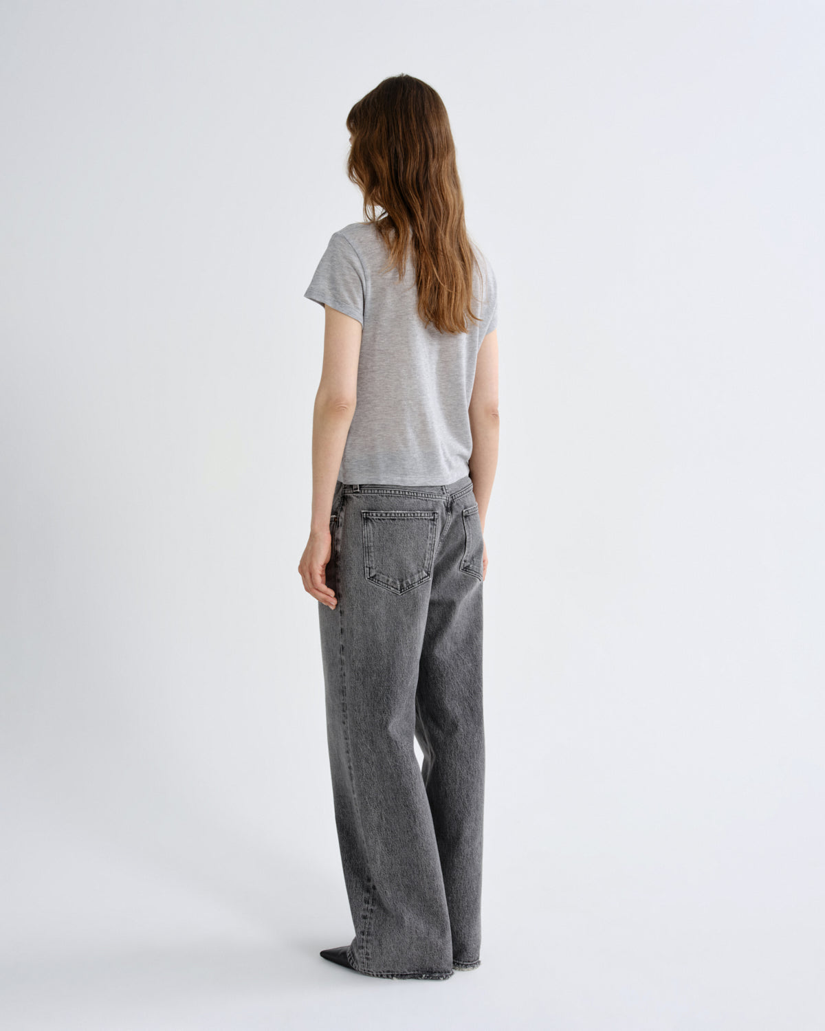 A woman viewed from the back wearing a light gray short-sleeve t-shirt and dark gray wide-leg jeans, standing against a plain light gray studio background.