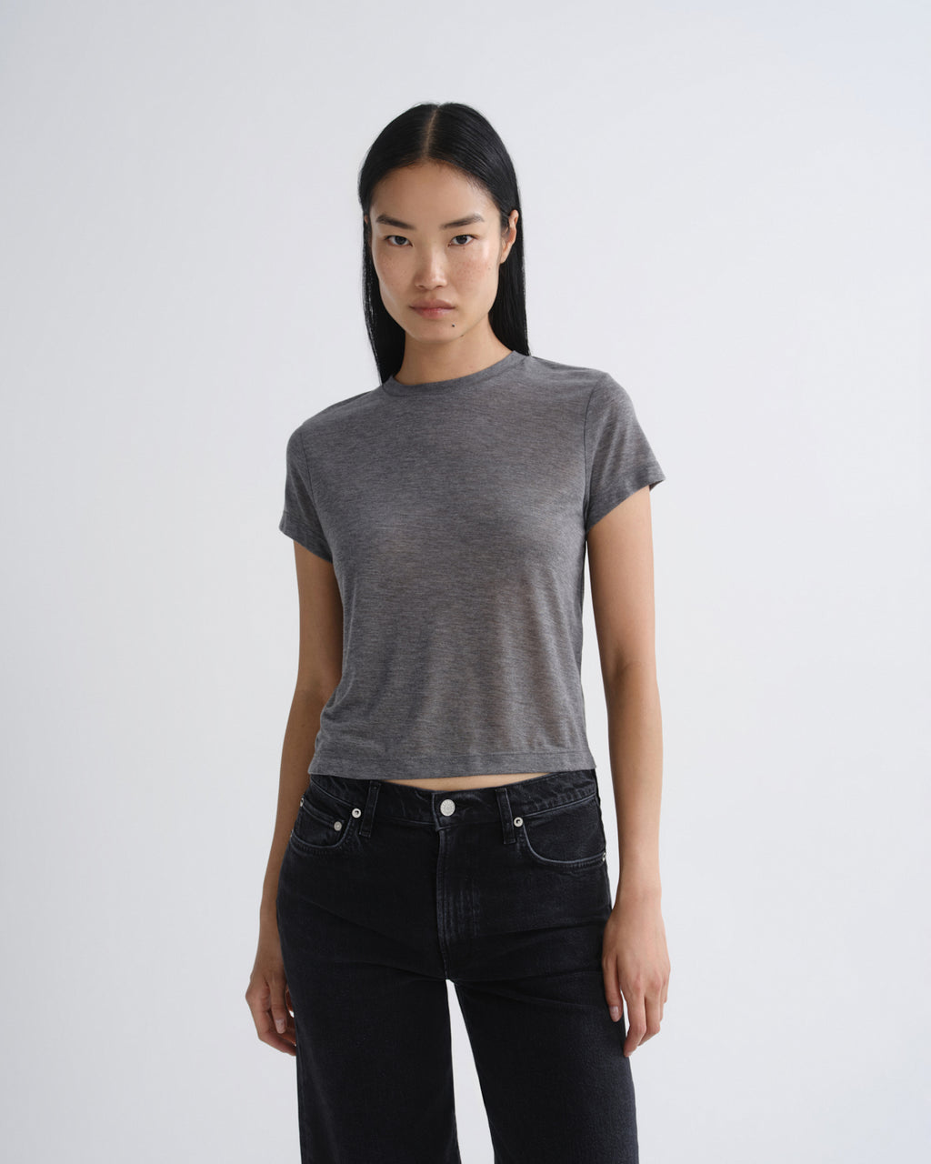 A woman modeling a short-sleeve, crew-neck gray crop top paired with black jeans, standing against a light gray studio background.