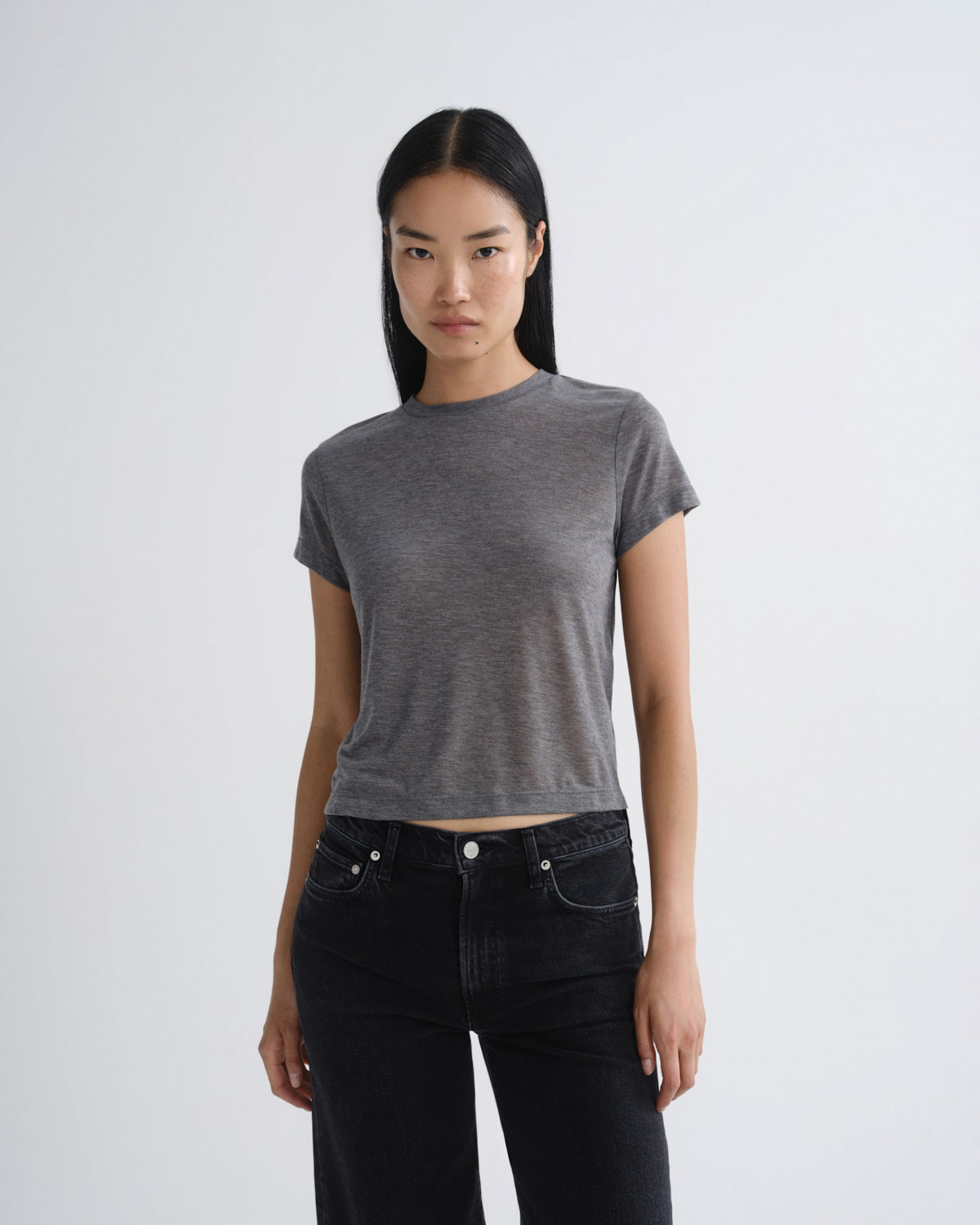 A woman modeling a short-sleeve, crew-neck gray crop top paired with black jeans, standing against a light gray studio background.
