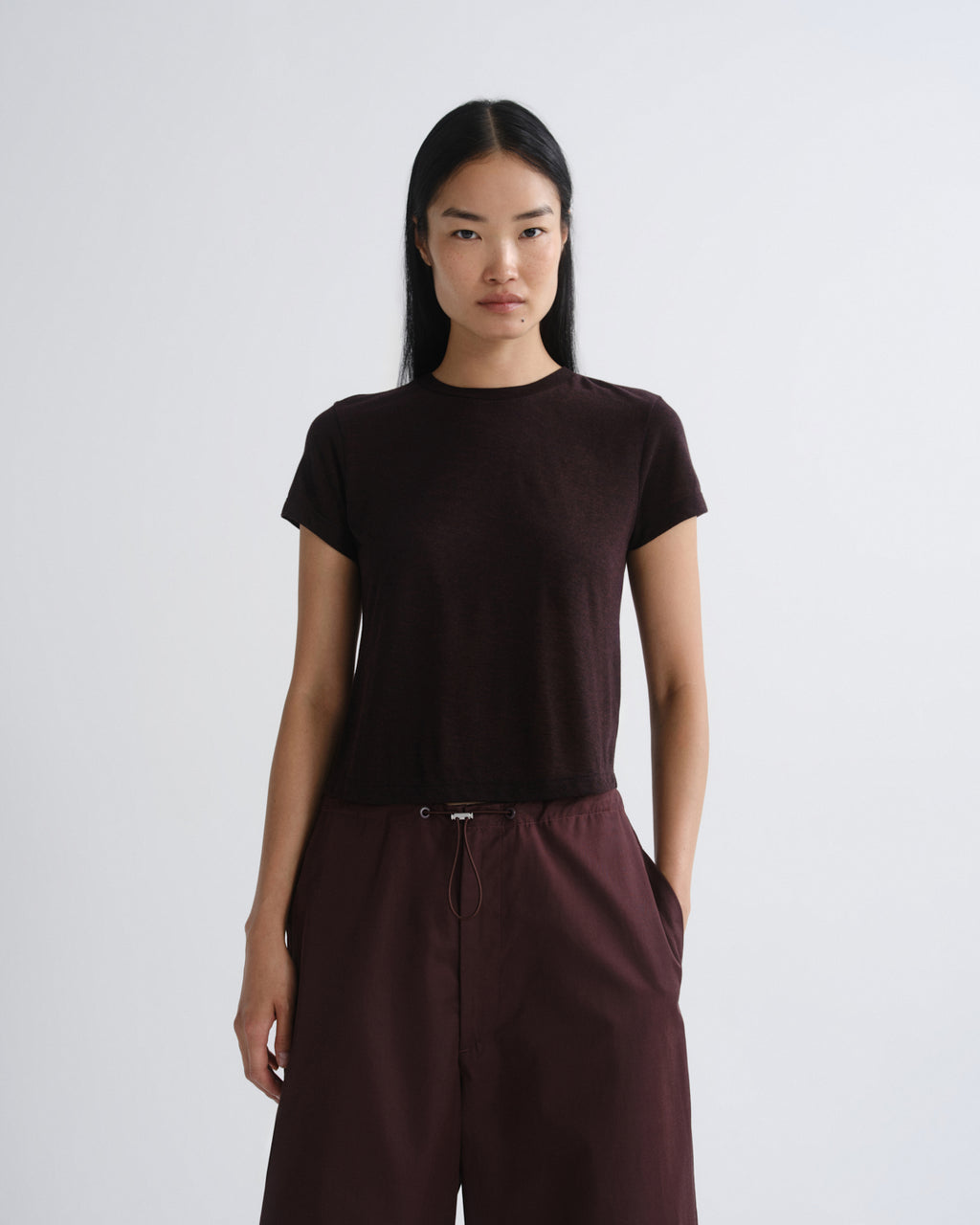 A model wearing a dark brown short-sleeve crewneck t-shirt and matching dark brown wide-leg trousers, standing against a light gray studio background.