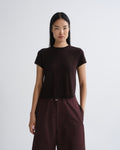 A model wearing a dark brown short-sleeve crewneck t-shirt and matching dark brown wide-leg trousers, standing against a light gray studio background.
