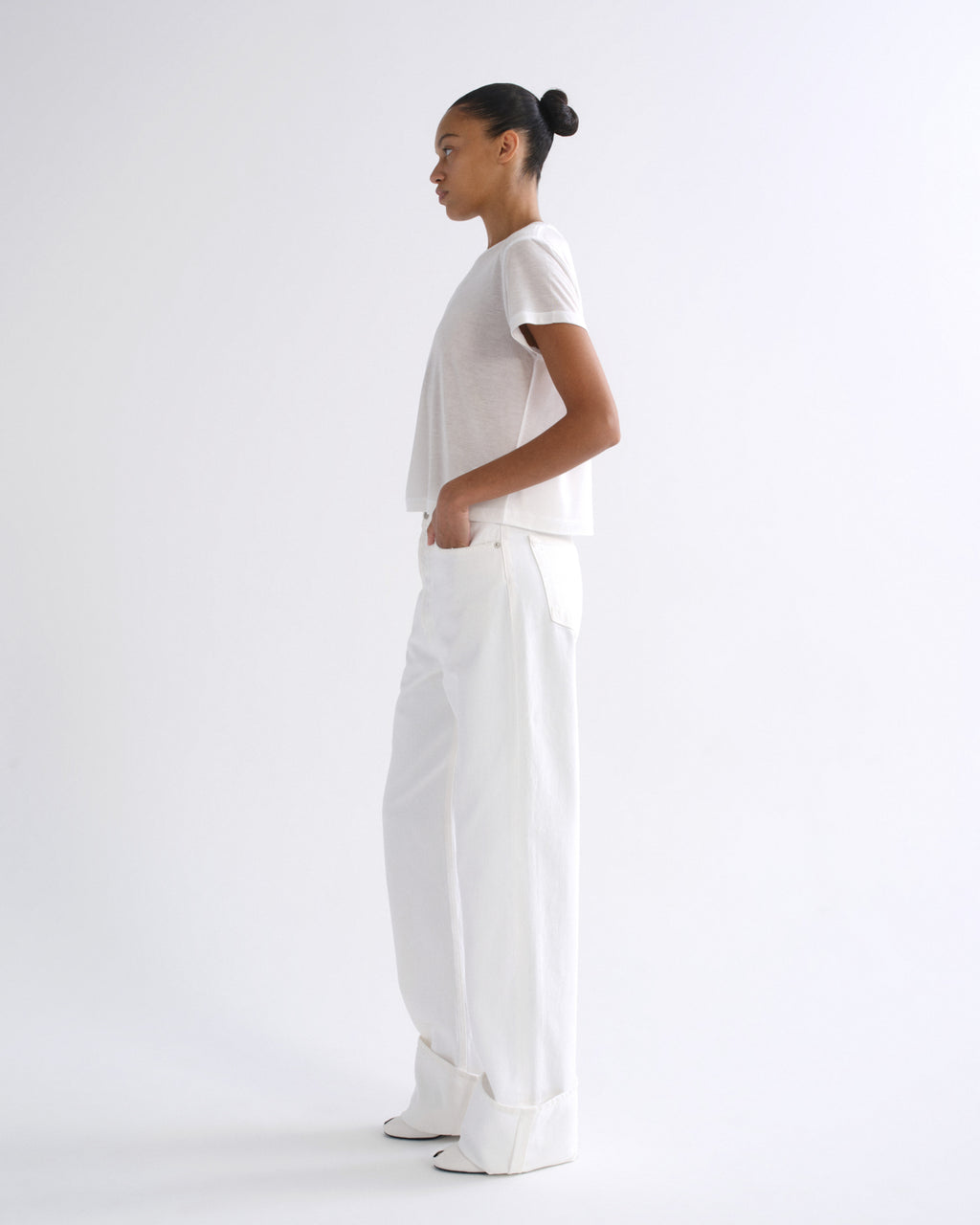 Woman wearing a white short sleeve crewneck tee with white straight leg pants viewed from the side on a grey background.