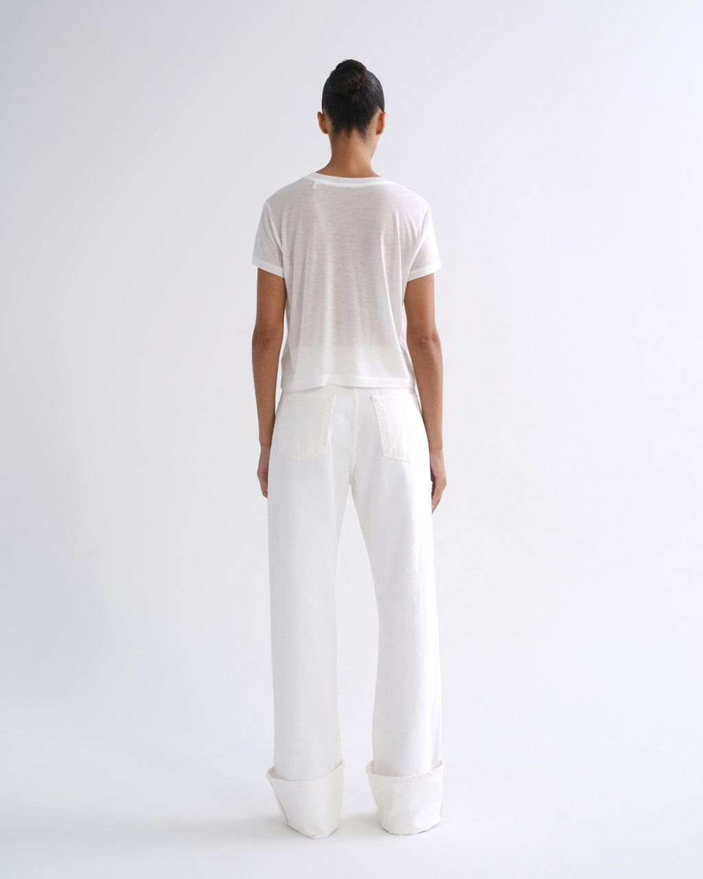 Woman wearing a white short sleeve crewneck tee with white straight leg pants viewed from the back on a grey background.