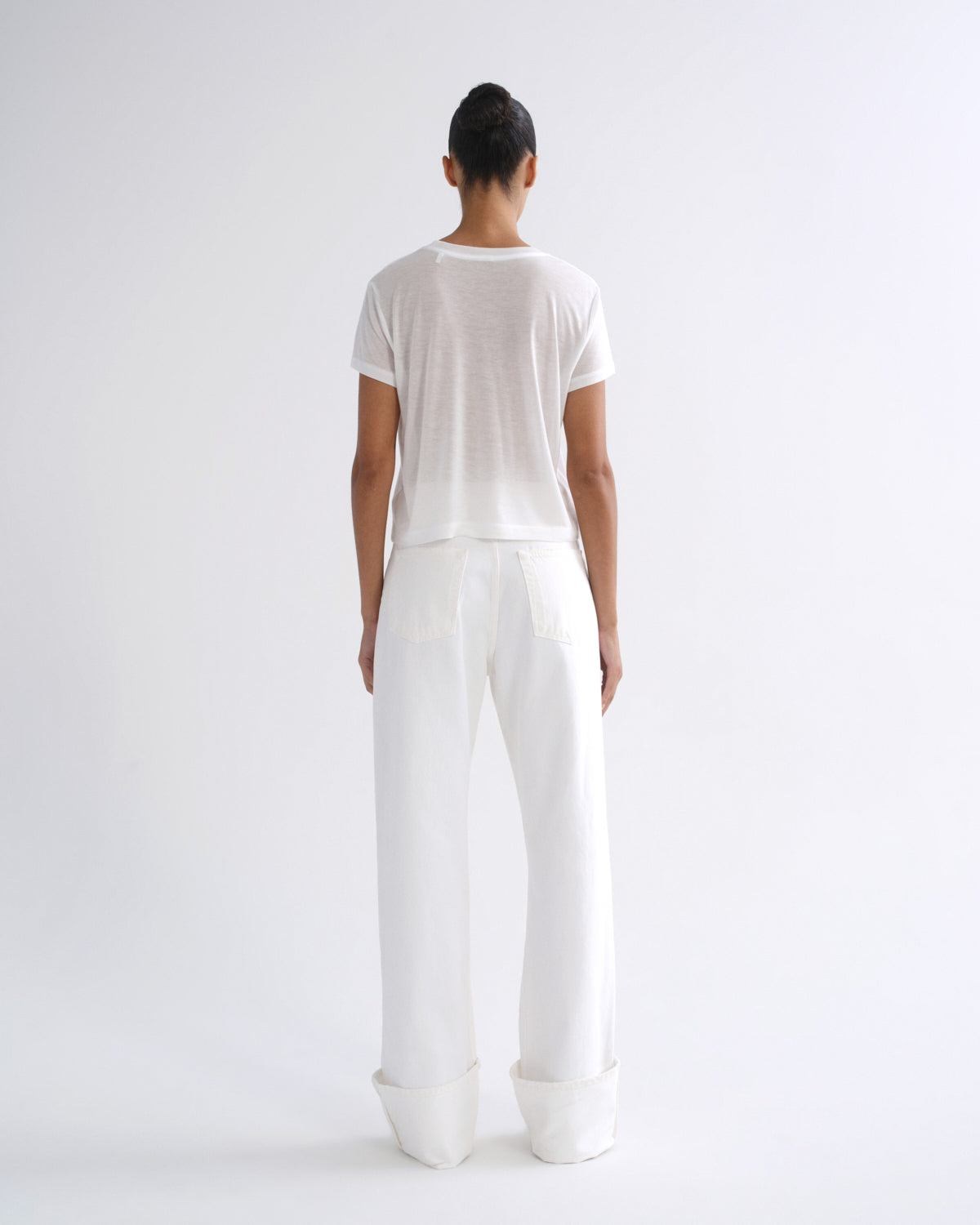 Woman wearing a white short sleeve crewneck tee with white straight leg pants viewed from the back on a grey background.