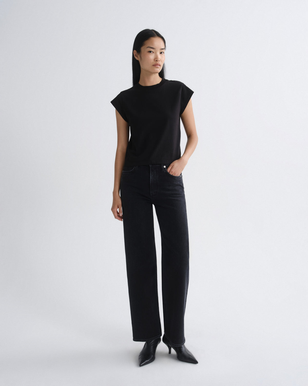 A model stands against a light gray studio background wearing a black short-sleeve crewneck top, black wide-leg jeans, and black pointed-toe heels.