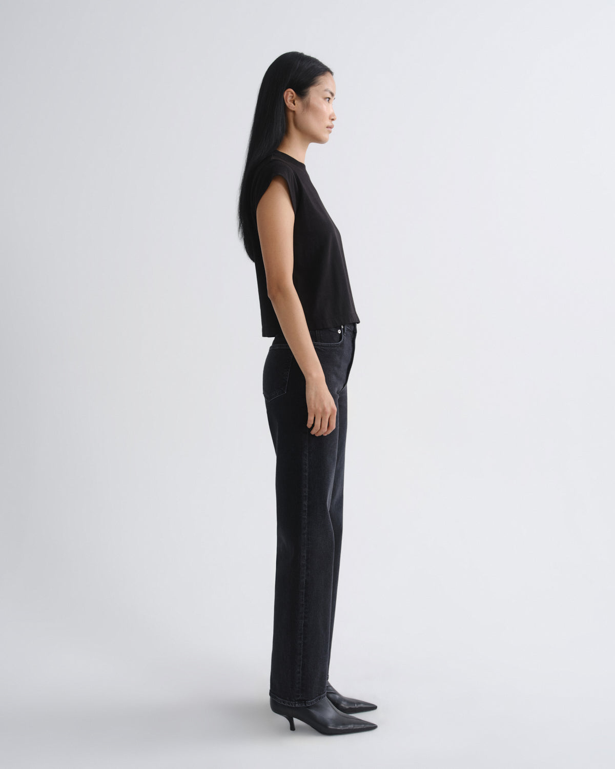 A woman stands in profile wearing a black sleeveless top, black wide-leg jeans, and black pointed-toe heels against a plain light gray studio background.