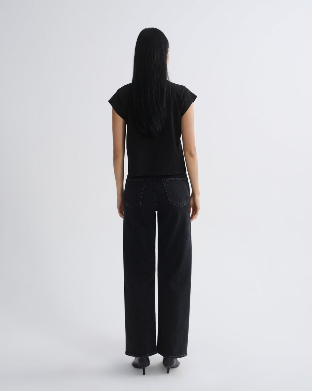 A woman viewed from the back wearing a black short-sleeve top and black wide-leg pants against a plain light gray studio background.