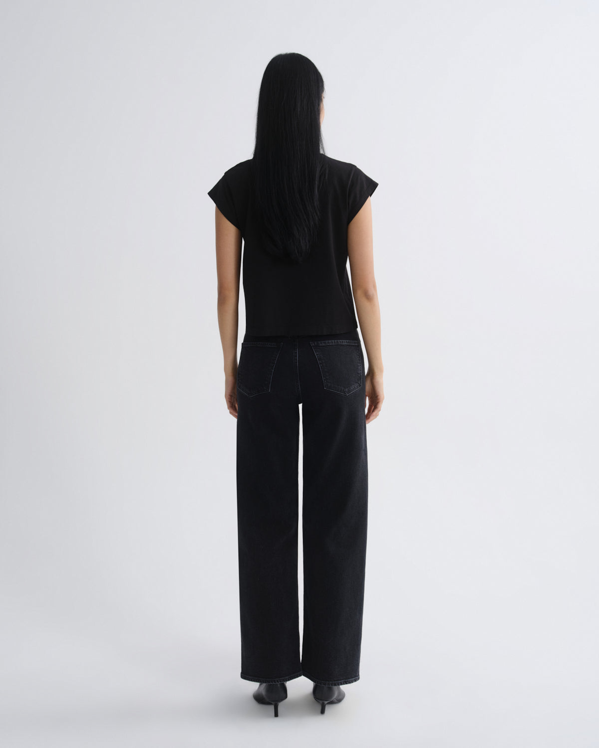A woman viewed from the back wearing a black short-sleeve top and black wide-leg pants against a plain light gray studio background.