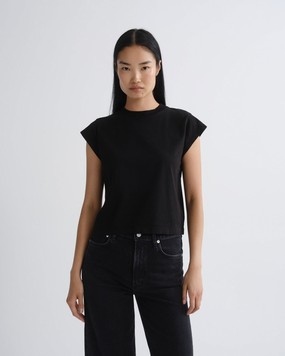 A woman modeling a black short-sleeve crop top with a crew neck, paired with black jeans, against a plain light gray studio background.