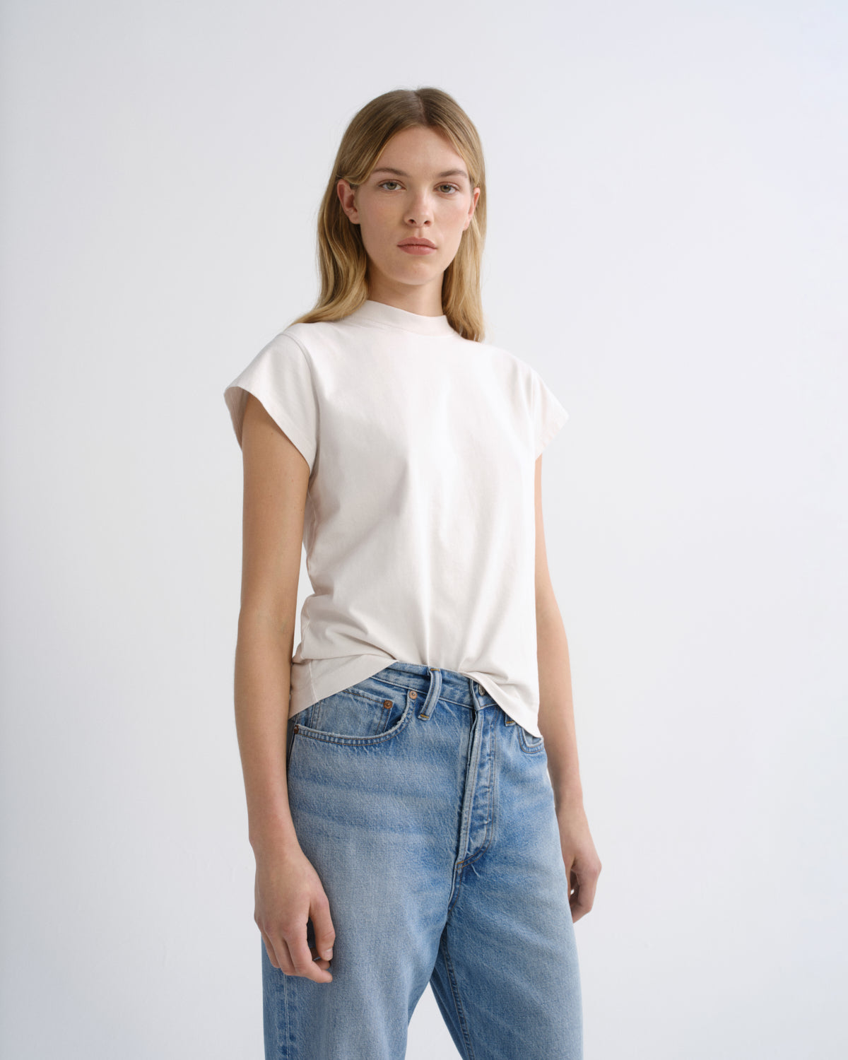 A woman modeling a short-sleeve crewneck t-shirt in off-white, tucked into light-wash blue jeans, against a plain light gray studio background.
