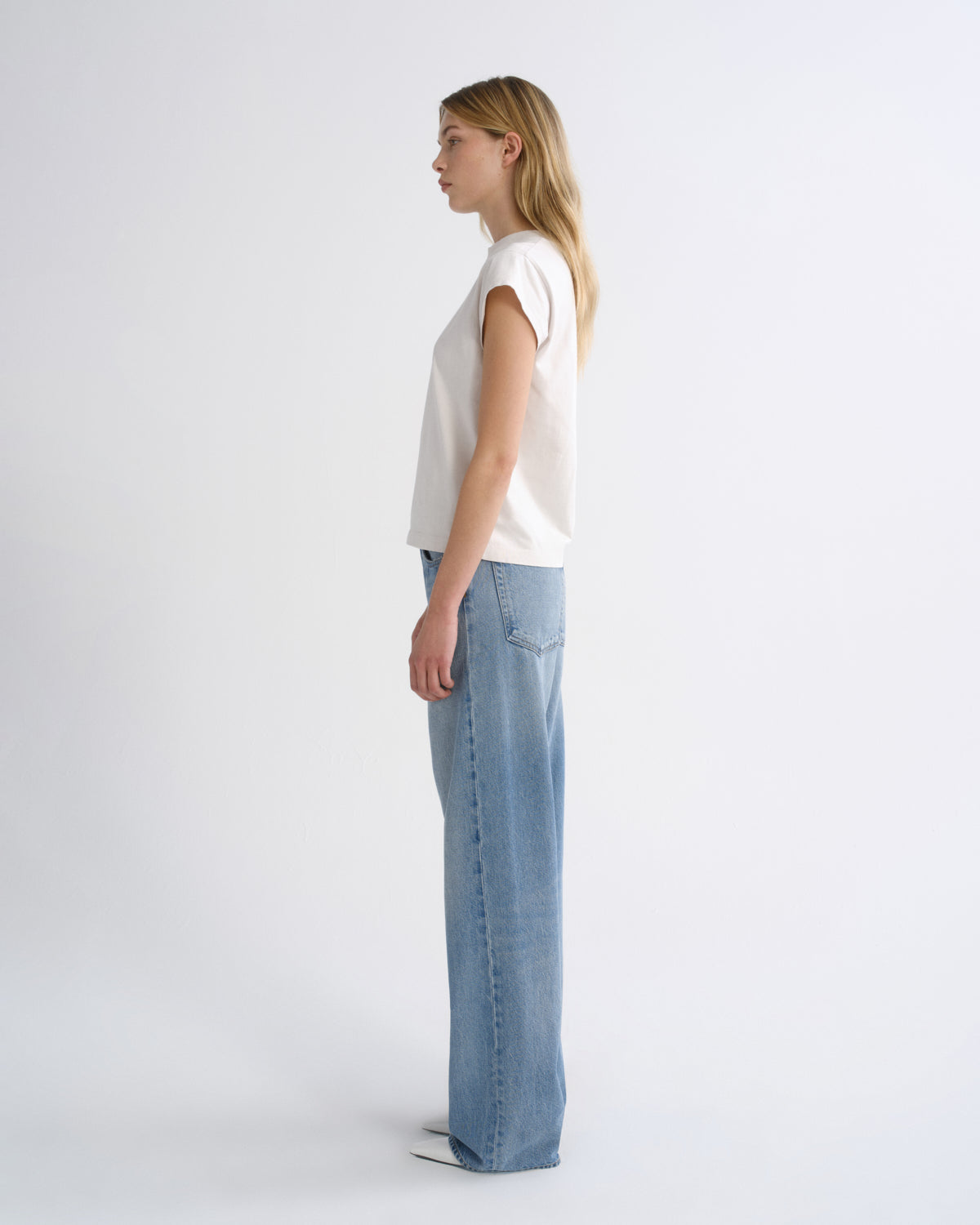 A woman stands in profile wearing a white short-sleeve top and light blue wide-leg jeans against a plain light gray studio background.