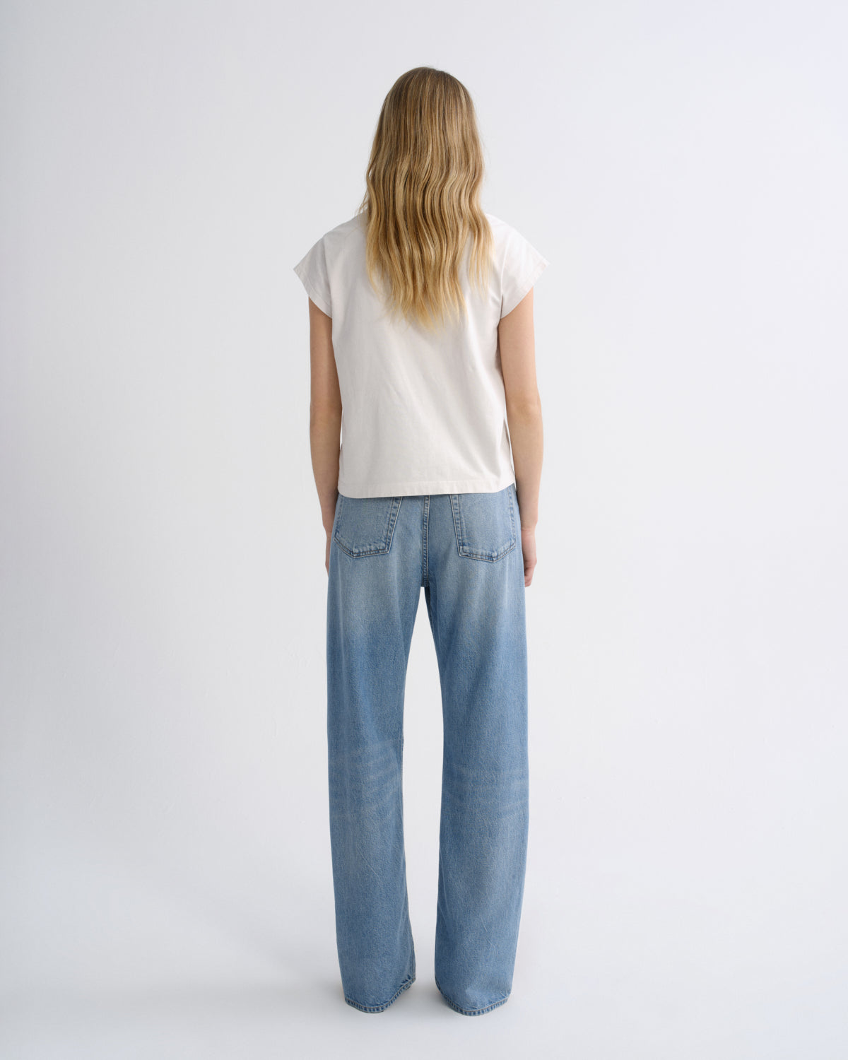 A woman viewed from behind wearing a white short-sleeve t-shirt and light blue wide-leg jeans, standing against a plain white studio background.