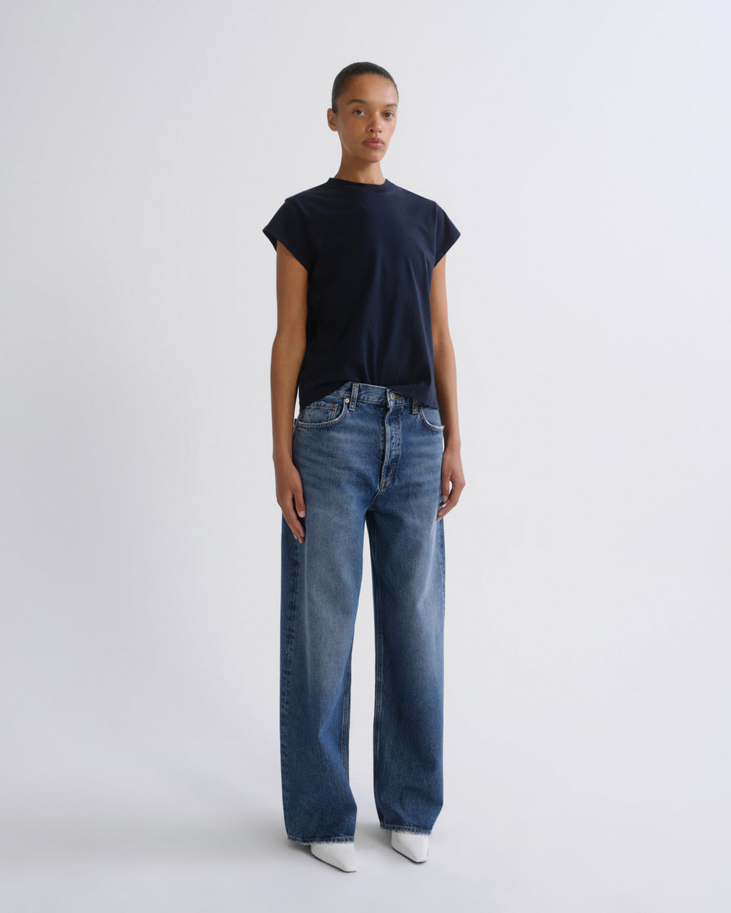 A model stands against a light gray studio background wearing a dark navy short-sleeve t-shirt, blue wide-leg jeans, and white pointed shoes.