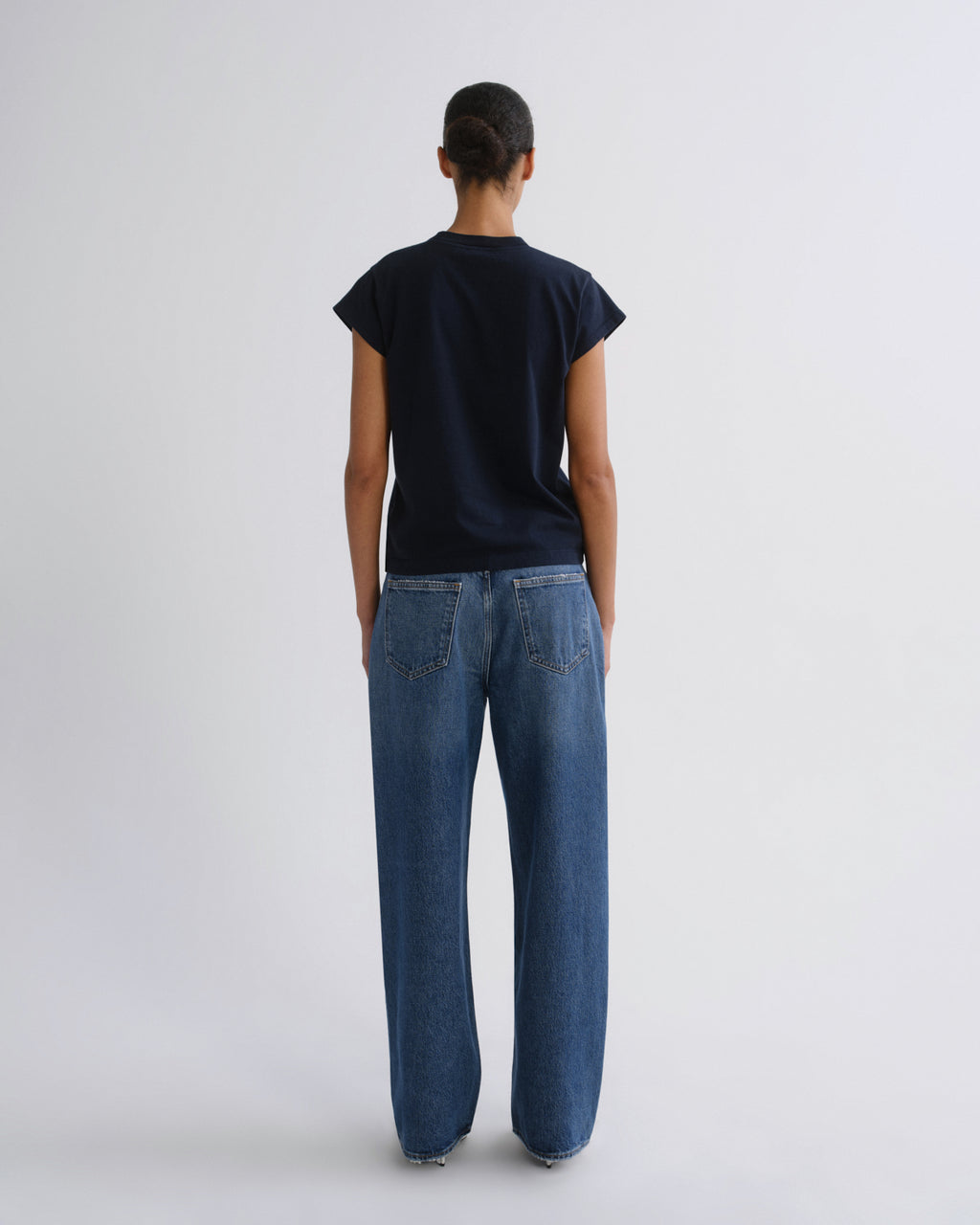 A model viewed from behind wearing a dark navy blue short-sleeve t-shirt and wide-leg blue denim jeans, standing against a light gray studio background.
