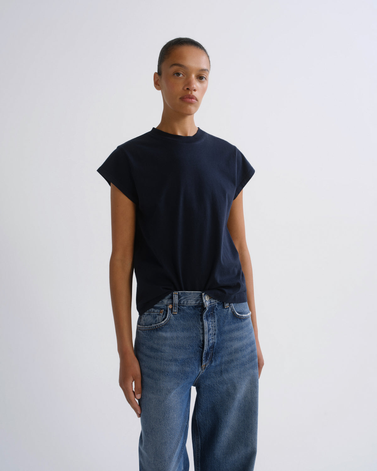 A model wearing a dark navy blue short-sleeve crewneck t-shirt and blue jeans, standing against a plain light gray studio background.