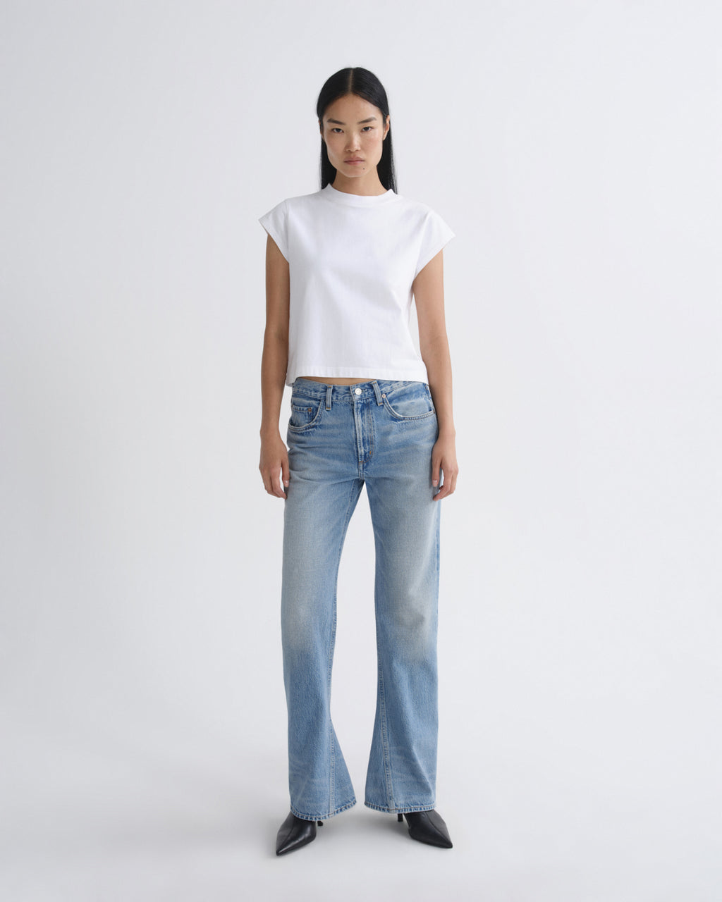 A model stands against a light gray studio background wearing a white short-sleeve cropped T-shirt, light-wash blue jeans, and black pointed shoes.