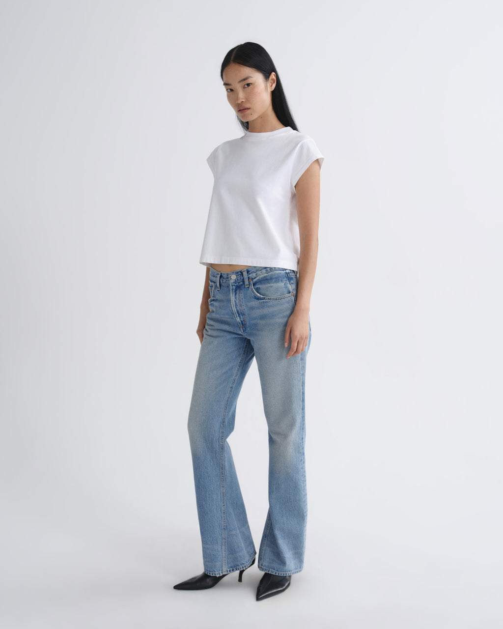 A model wearing a white short-sleeve crop top, light-wash blue flare jeans, and black pointed-toe heels, standing against a plain light gray studio background.