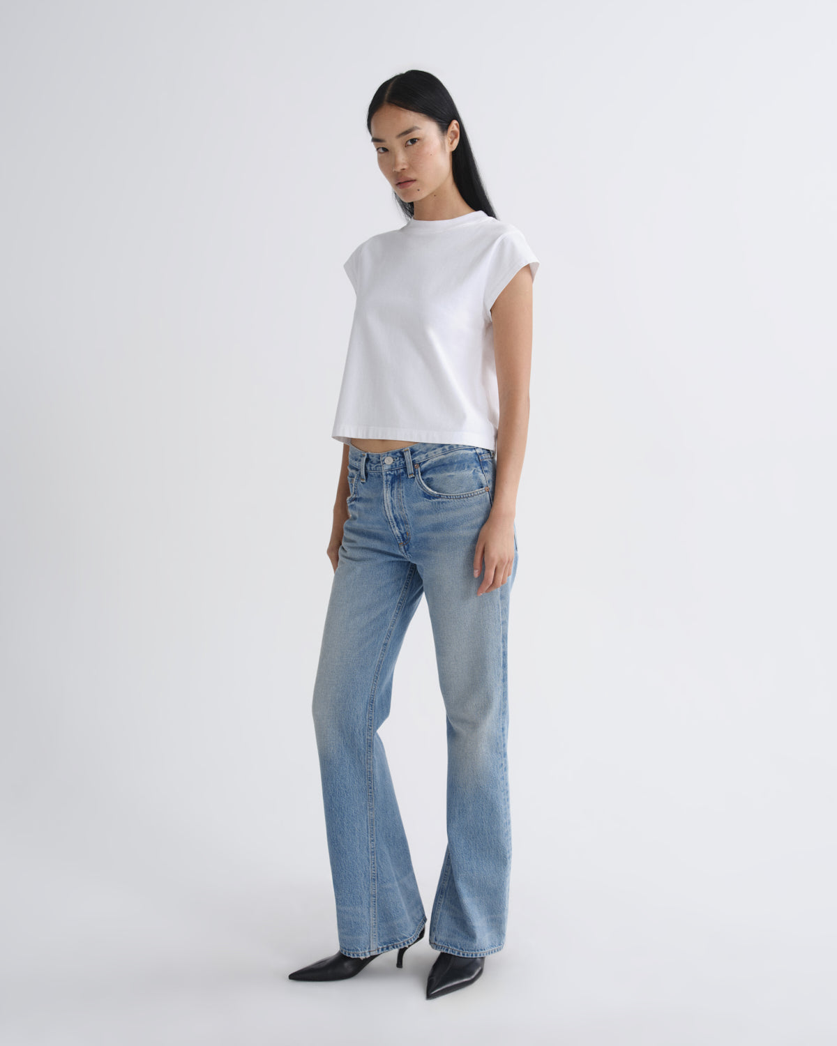 A model wearing a white short-sleeve crop top, light-wash blue flare jeans, and black pointed-toe heels, standing against a plain light gray studio background.