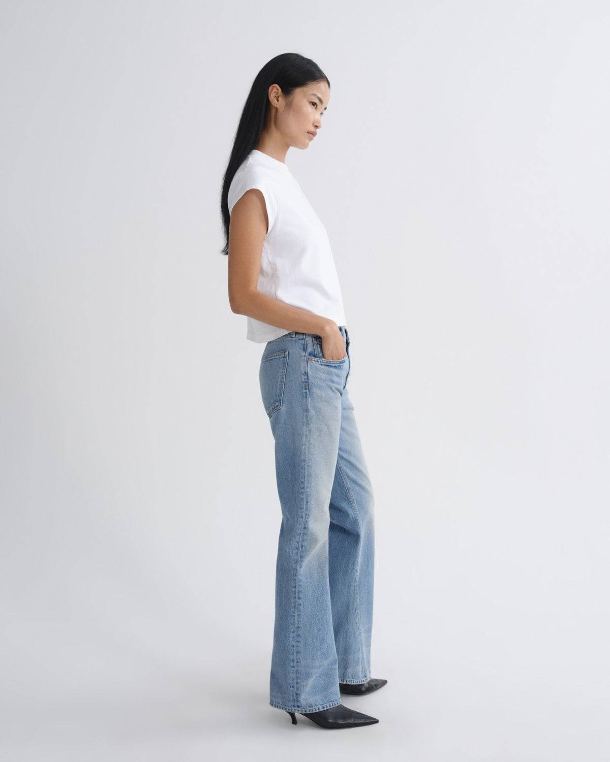 A woman in a white sleeveless top and light blue wide-leg jeans stands in profile against a plain light gray studio background.