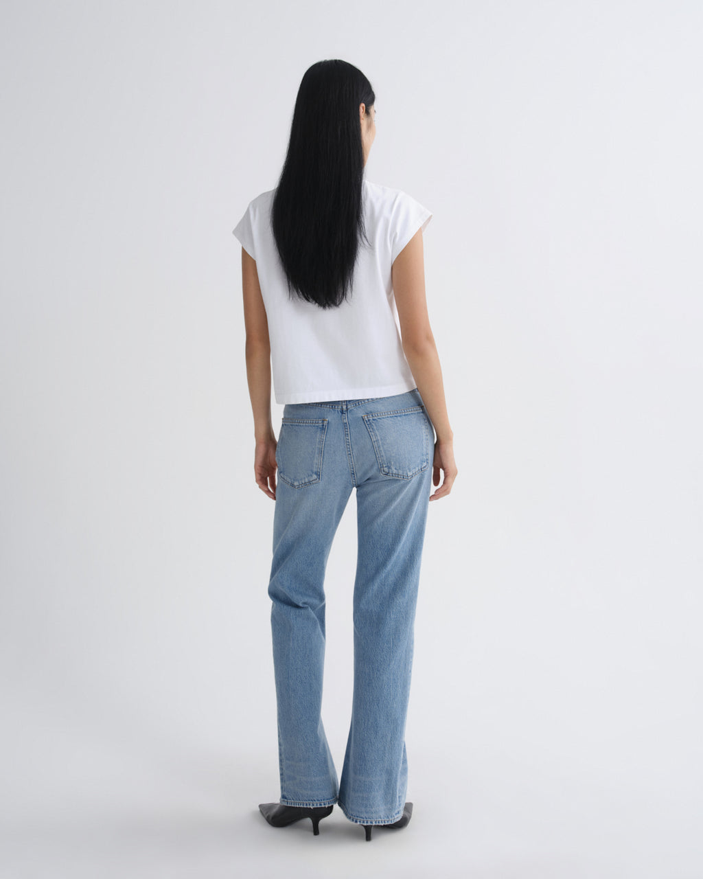 A woman viewed from behind wearing a white short-sleeve top, light blue straight-leg jeans, and black pointed-toe heels against a plain light gray studio background.