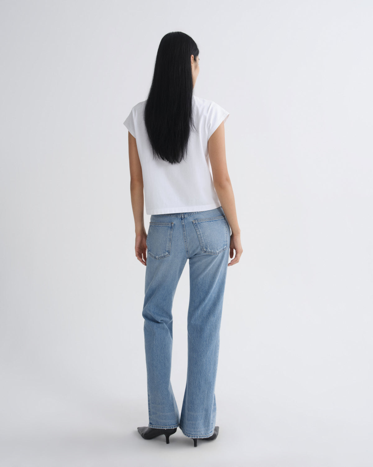 A woman viewed from behind wearing a white short-sleeve top, light blue straight-leg jeans, and black pointed-toe heels against a plain light gray studio background.