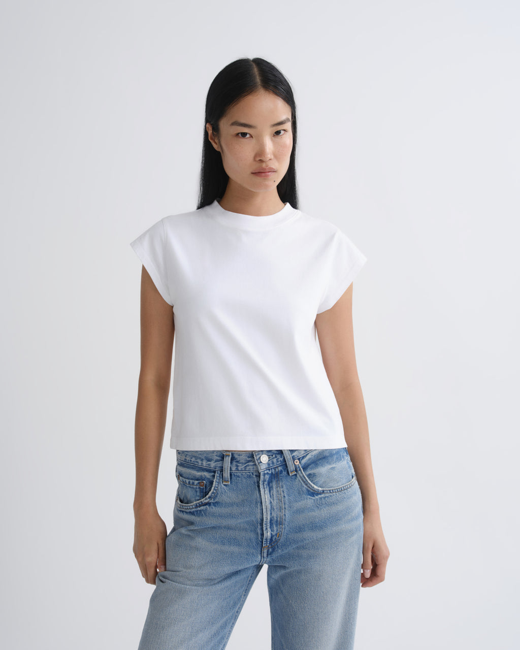 A woman modeling a white short-sleeve crewneck crop top paired with light-wash blue jeans, photographed against a clean light gray studio background.