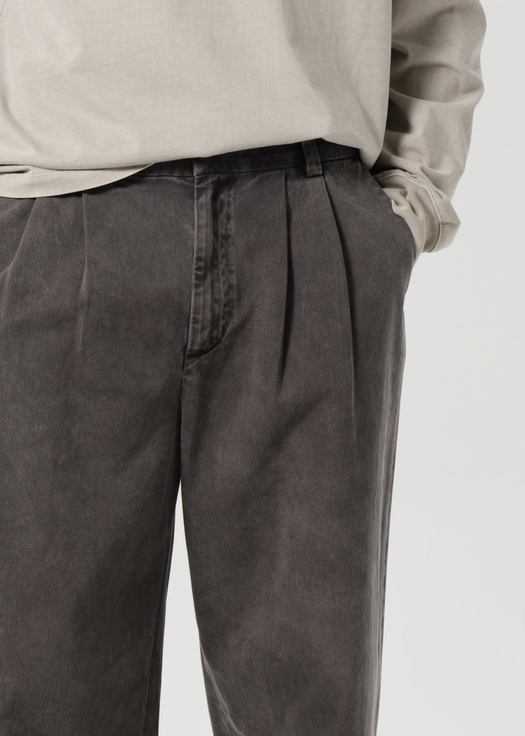 Person wearing dark pleated pants and a light-colored shirt on a plain background