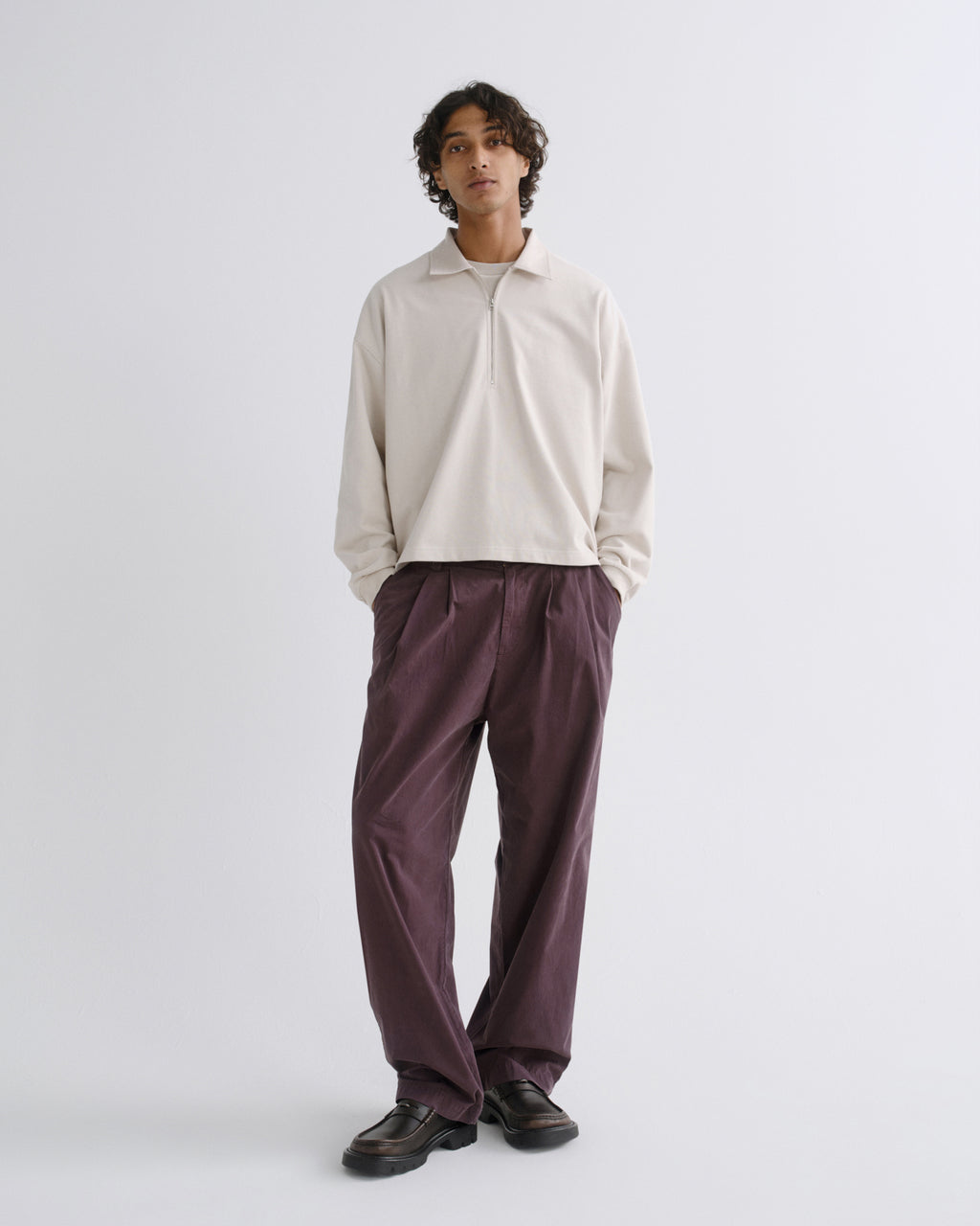 A model stands against a light gray studio background wearing a cream long-sleeve polo shirt and dark purple wide-leg trousers with dark brown leather shoes.