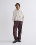 A model stands against a light gray studio background wearing a cream long-sleeve polo shirt and dark purple wide-leg trousers with dark brown leather shoes.