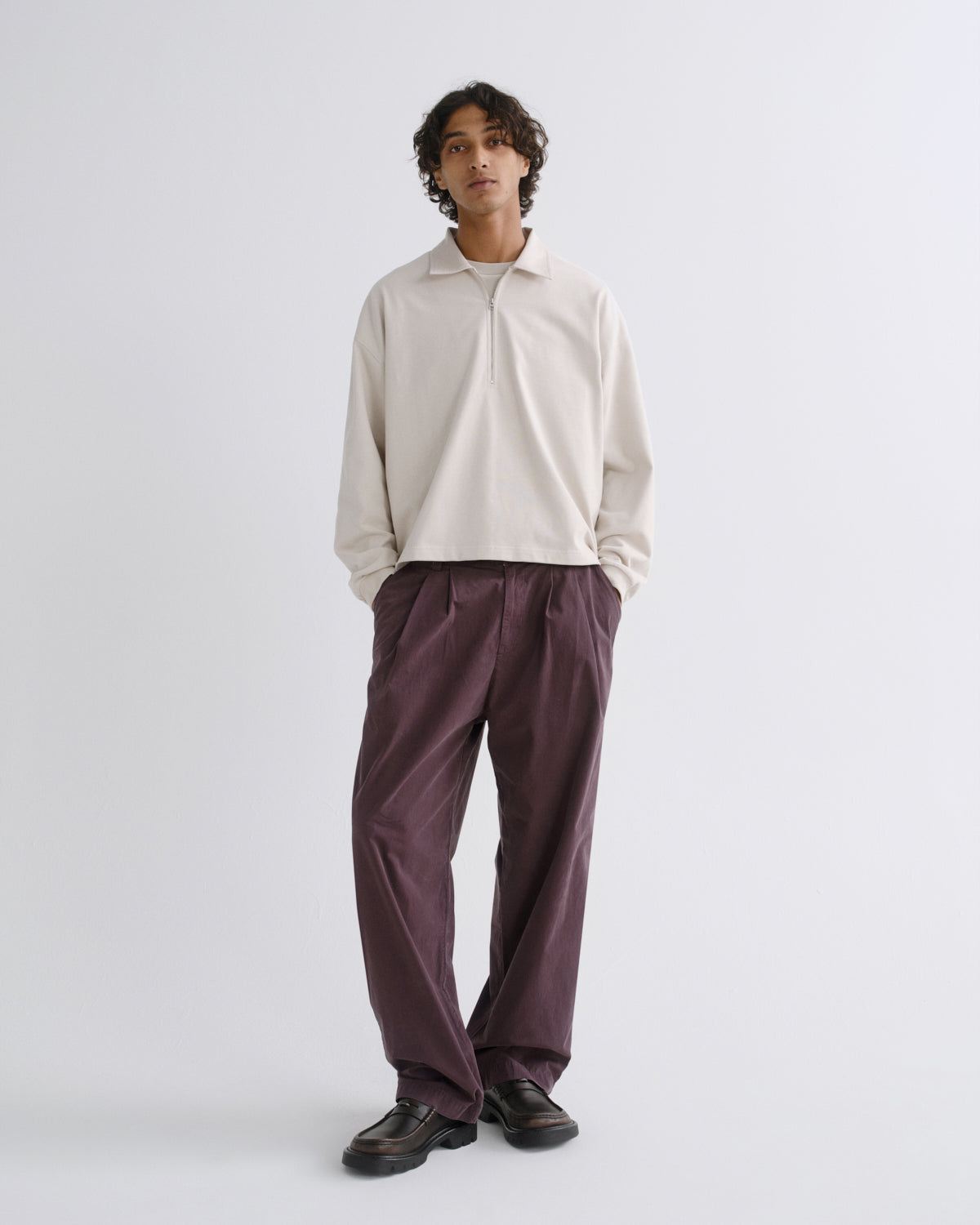 A model stands against a light gray studio background wearing a cream long-sleeve polo shirt and dark purple wide-leg trousers with dark brown leather shoes.
