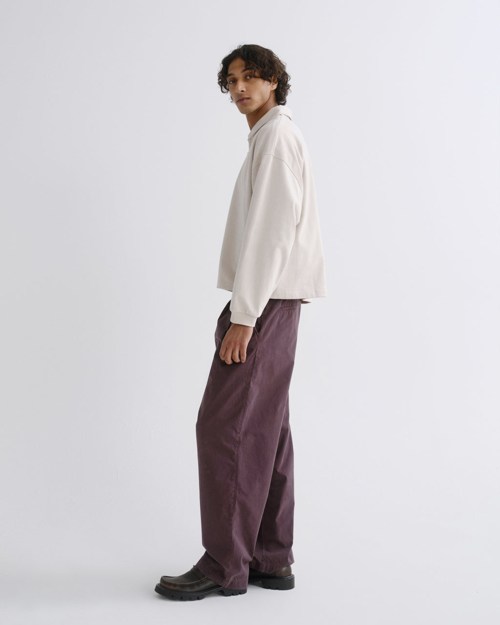 A model wearing a cream long-sleeve shirt and wide-leg mauve trousers, standing in profile against a plain light gray studio background.