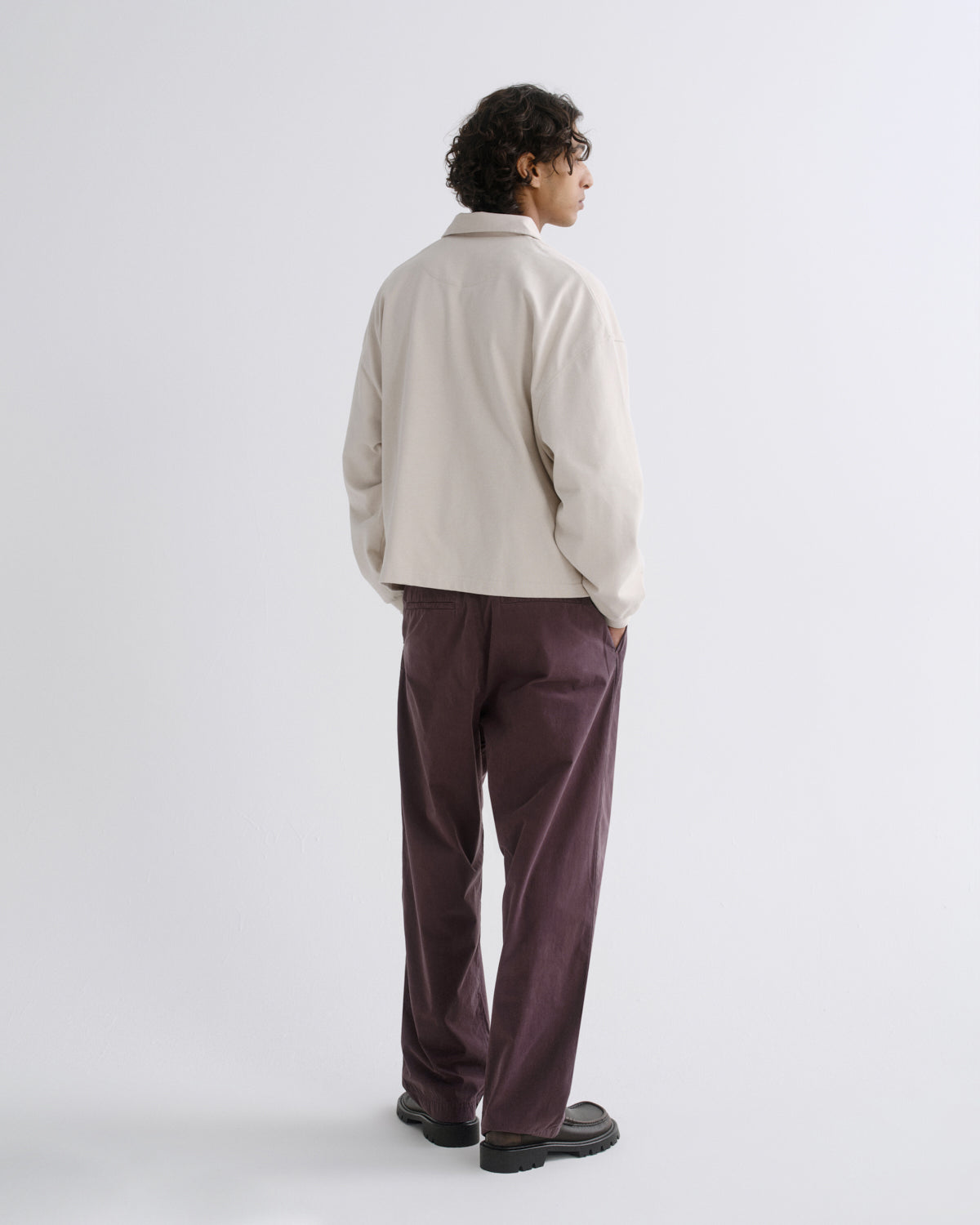 A man is shown from the back wearing a light cream-colored jacket and dark purple trousers, standing against a plain light gray studio background.