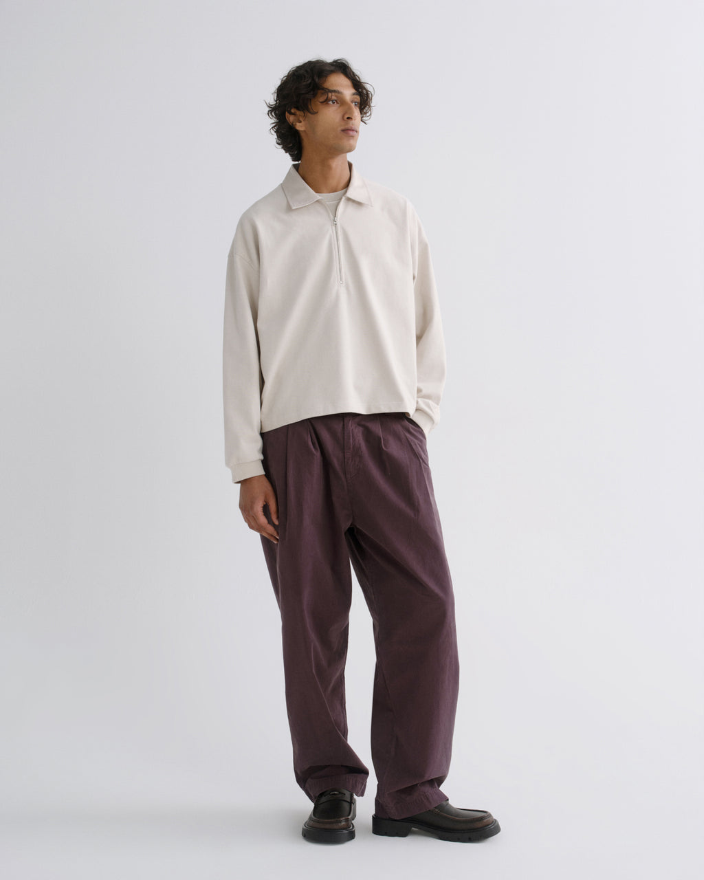 A model stands in a studio wearing a cream long-sleeve polo shirt and dark plum wide-leg trousers, paired with black shoes, against a light gray background.