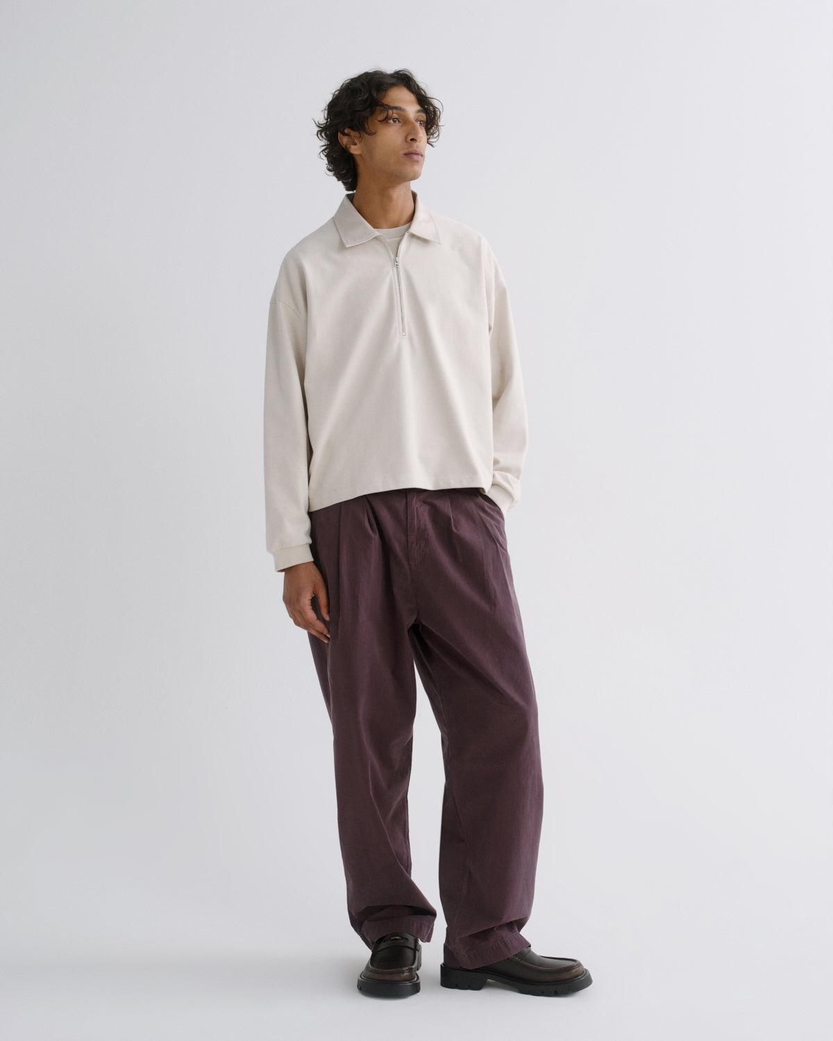 A model stands in a studio wearing a cream long-sleeve polo shirt and dark plum wide-leg trousers, paired with black shoes, against a light gray background.