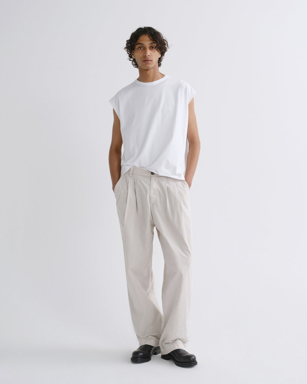 A model stands against a light gray studio background wearing a white sleeveless t-shirt, cream pleated trousers, and black loafers.